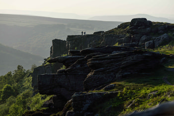 Peak district edge This landscape photograph captures Curbar Edge in the Peak District, located in Derbyshire, United Kingdom, during a spring evening. The main subject of the image is the prominent rocky escarpment of Curbar Edge, which rises above the surrounding rural terrain and showcases the dramatic geological formations typical of the Peak District. In the distance, two figures stand atop the rocks, emphasizing the scale and natural beauty of the area. The scene is bathed in soft evening light, casting long shadows across the rugged landscape and highlighting the lush greenery of spring nature below. The rolling hills and layered cliffs demonstrate the unique character of this rural region in the United Kingdom.